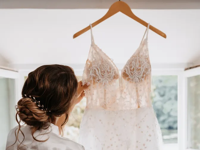 A bride in a white robe reaching up to her wedding dress where it hangs in a bright window.