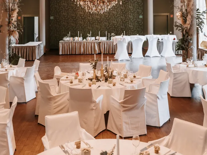 A wedding reception hall with a large chandelier, round tables in white linens and a long top table.