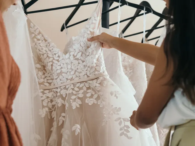 A woman's hand lifting the strap of an embroidered wedding dress from a rail of gowns in a boutique.