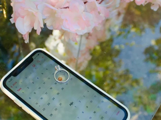 An engagement ring resting on a phone showing a calendar, surrounded by soft pink blossom, symbolising choosing a wedding date.
