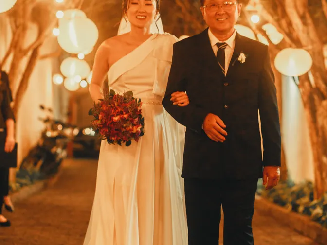 A smiling bride walking arm in arm with her father beneath strings of glowing paper lanterns at a night-time wedding.