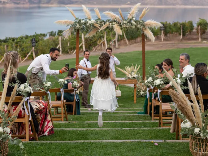 A young flower girl in a white dress running down a grass aisle towards a floral arch at an outdoor wedding, guests watching from wooden chairs.