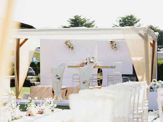 An elegant outdoor wedding reception set up under a white marquee, with draped fabric, gold chairs and pastel floral arrangements.