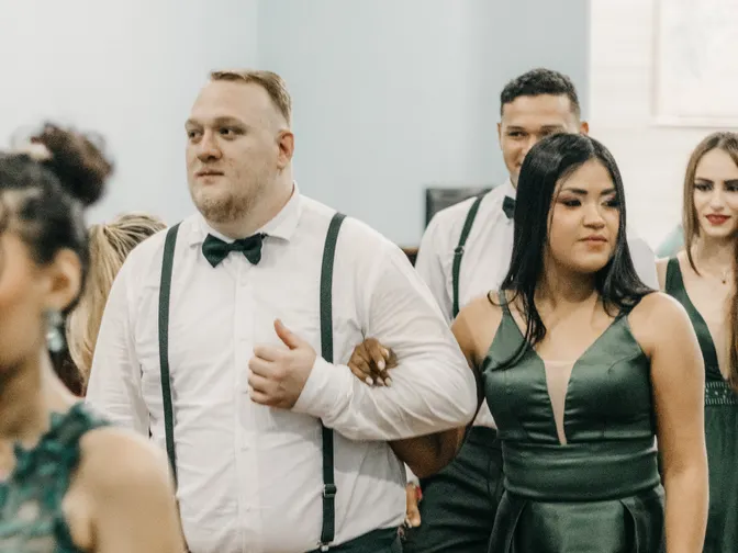 Members of a wedding party in dark green dresses and braces walking arm in arm down the aisle of a church.