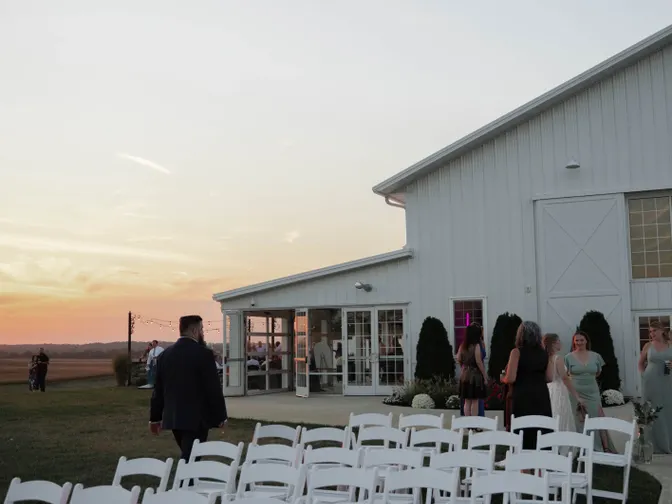 Rows of white folding chairs set for an outdoor ceremony beside a white barn venue as the sun sets.