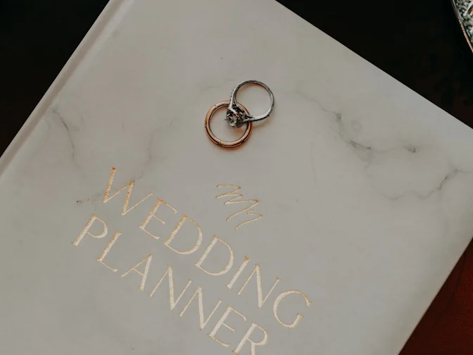 A marble-effect wedding planner book with two rings resting on top, beside a tray of pale pink blossom.